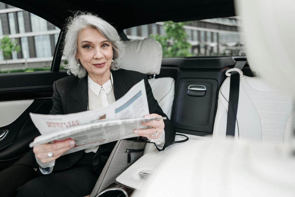 Senior businesswoman reading a newspaper while seated in a car, smiling confidently.